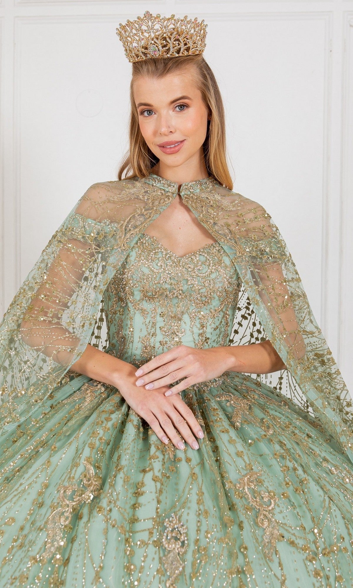 A woman wearing a gold crown and the Quinceanera Dress 4201 by Cinderella Couture—an ornate light green ball gown with gold embellishments—stands against a plain white background.